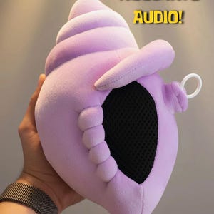 May include: A plush lavender seashell-shaped object with a black mesh opening on the front, a small white ring attached to the side, and the text 100% ACCURATE AUDIO! printed in yellow above the shell. The soft fabric has a smooth texture.