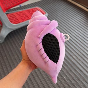 May include: A soft, plush seashell toy in a light purple color. The shell features a spiral-shaped top, a textured black mesh opening on the side, and a small white plastic ring attached to the right side of the structure for hanging or carrying.
