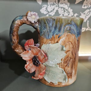 May include: A ceramic mug with a textured wood grain finish in shades of brown, blue, and green. The handle is shaped like a tree branch with a small flower. A large, sculpted orange flower with a dark centre and pale green leaves adorns the side.