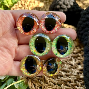 May include: Six glass doll eyes in various colours, including red, green, and gold, with black pupils. The eyes have glitter accents and are round. These eyes are for crafts and doll making.