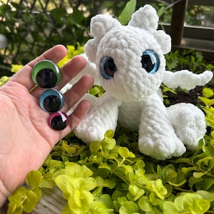 Può includere: Un peluche di drago bianco all'uncinetto con grandi occhi blu, ali e piccole orecchie. Tre occhi di vetro in verde, blu e rosa sono tenuti vicino. Il drago è seduto su un letto di piante verdi.