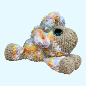 Peut inclure: Un jouet en peluche crocheté en forme d'agneau avec un visage et des sabots de couleur beige unie. Le corps et les oreilles présentent un motif de fil panaché dans des tons pastel de rose, jaune, bleu et blanc. Il a de grands yeux sombres.