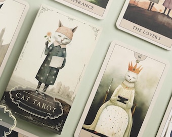 Könnte beinhalten: Ein Set Tarotkarten mit Illustrationen von Katzen. Die Karten befinden sich in einer weißen Schachtel mit dem Text "Cat Tarot" auf der Vorderseite. Die Karten liegen auf einer hellgrünen Oberfläche. Die Karten beinhalten "The Lovers", "Temperance", "The Empress" und "Cat Tarot".