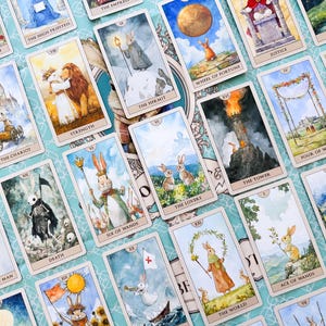 K&ouml;nnte beinhalten: Ein Set Tarotkarten mit fantasievollen Illustrationen von Kaninchen in verschiedenen Szenen. Die Karten zeigen Titel wie "The Lovers", "The Hermit" und "The Tower" auf einem hellblauen Hintergrund.