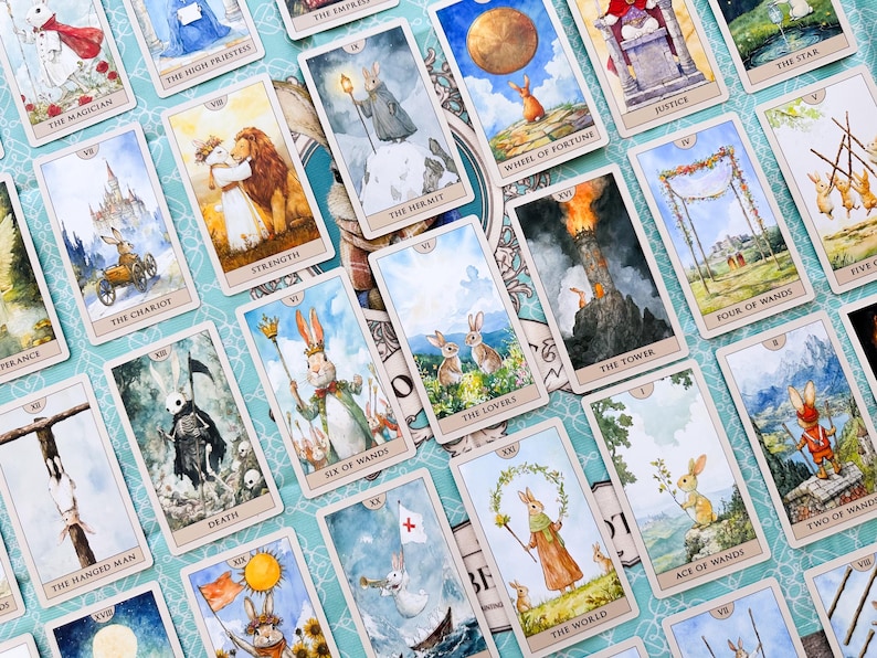 K&ouml;nnte beinhalten: Ein Set Tarotkarten mit fantasievollen Illustrationen von Kaninchen in verschiedenen Szenen. Die Karten zeigen Titel wie "The Lovers", "The Hermit" und "The Tower" auf einem hellblauen Hintergrund.