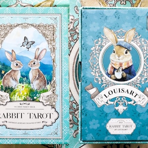 K&ouml;nnte beinhalten: Ein Rabbit Tarot Kartenspiel in einer t&uuml;rkisfarbenen Box. Die Vorderseite der Box zeigt eine Kaninchen-Illustration mit dem Text "RABBIT TAROT" und "78 CARD TAROT DECK". Die Box-Art beinhaltet einen Schmetterling und zwei Kaninchen. Die Box hat auch den Text "LOUISART".