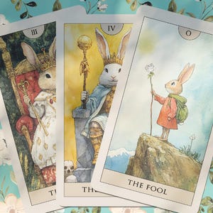 K&ouml;nnte beinhalten: Drei Tarotkarten mit Kaninchen-Illustrationen. Die Karten zeigen ein Kaninchen mit Krone, ein Kaninchen mit Stab und ein Kaninchen mit Blume. Die Karten sind vor einem floralen Hintergrund angeordnet.