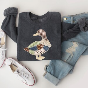 May include: A dark gray sweatshirt featuring a patchwork duck design with various patterns and lace trim. The outfit includes light blue denim jeans and white Converse sneakers. A casual, comfortable ensemble.