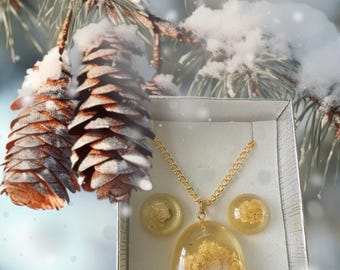 Puede incluir: Un conjunto de collar y pendientes de oro con un colgante de resina transparente y pendientes con flores blancas secas. Las joyas se muestran en una caja blanca sobre un fondo nevado.