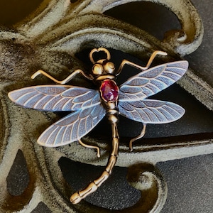 May include: A detailed dragonfly pendant with a purple gemstone in the center. The dragonfly is made of metal with a silver finish and has intricate details on its wings.
