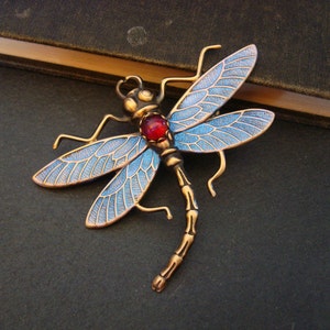 May include: A dragonfly brooch featuring a metallic bronze-toned body and wings with blue enamel detailing. The insect has a round, translucent red stone set in the center of its thorax, with intricate wing veins and a segmented tail design.