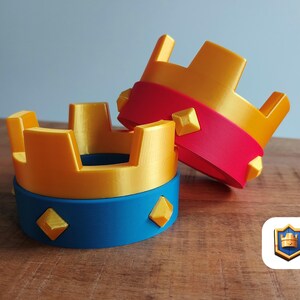 May include: Two 3D-printed crowns. One is upright, the other is leaning. The crowns are gold with red and blue accents. Each crown has a blue base with gold diamond shapes. The background is a light blue wall and a wooden table.
