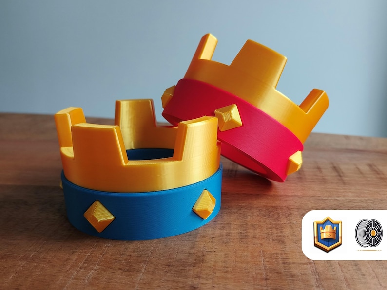 May include: Two 3D-printed crowns. One is upright, the other is leaning. The crowns are gold with red and blue accents. Each crown has a blue base with gold diamond shapes. The background is a light blue wall and a wooden table.