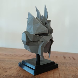 Puede incluir: Una escultura geométrica gris de un casco con rasgos angulares y afilados. El casco está montado sobre un soporte negro con una base cuadrada. La escultura tiene un diseño moderno y abstracto.