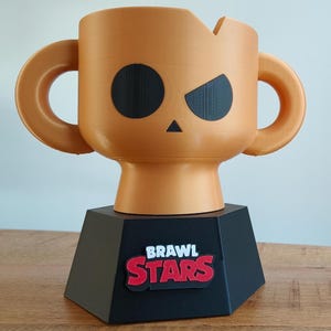 Op de afbeelding: Een 3D-geprinte trofee in de vorm van een personage uit het spel Brawl Stars. De oranje trofee heeft zwarte gezichtskenmerken en handvatten. Hij staat op een zwarte basis met het logo van het spel in rood en wit.