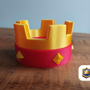 May include: A 3D-printed crown in yellow and red. The crown has a cylindrical base with yellow diamond-shaped accents. The top of the crown is yellow with rectangular spires. The object is on a wooden surface.