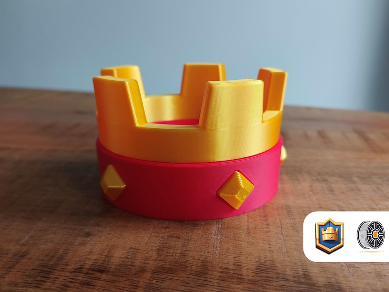 May include: A 3D-printed crown in yellow and red. The crown has a cylindrical base with yellow diamond-shaped accents. The top of the crown is yellow with rectangular spires. The object is on a wooden surface.