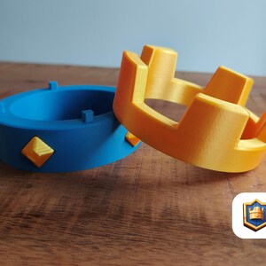 May include: Two 3D-printed crowns, one blue with yellow diamond accents and one yellow. The blue crown is a ring shape, while the yellow crown has a more traditional design. The items are on a wooden surface.