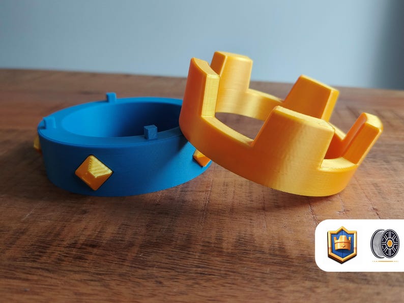 May include: Two 3D-printed crowns, one blue with yellow diamond accents and one yellow. The blue crown is a ring shape, while the yellow crown has a more traditional design. The items are on a wooden surface.