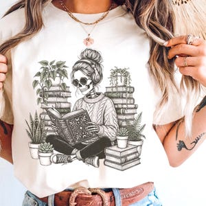 May include: Cream-colored t-shirt featuring a skeleton reading a book, surrounded by stacks of books and potted plants. The skeleton wears headphones and a cable-knit sweater. The design is in black and white.