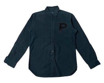 May include: Dark teal button-down shirt with long sleeves. Features a black patch with a white "P" on the left chest. The shirt has a classic collar and buttoned cuffs. The fabric appears to be a soft, woven material.
