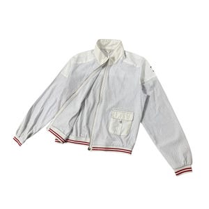 Puede incluir: Chaqueta blanca con cremallera y diseño perforado. Cuenta con cuello blanco, un pequeño bolsillo y puños y dobladillo a rayas rojas y blancas. La chaqueta tiene un estilo deportivo y atlético.