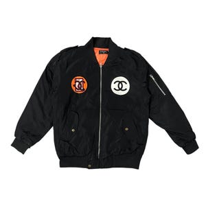 May include: Black bomber jacket with orange lining, featuring two circular patches with logos. The jacket has a zipper closure, two front pockets, and a zippered pocket on the left sleeve. The logos are in orange and white.