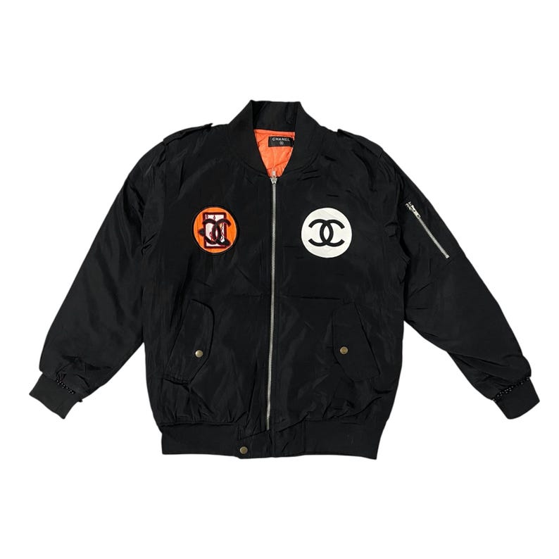 May include: Black bomber jacket with orange lining, featuring two circular patches with logos. The jacket has a zipper closure, two front pockets, and a zippered pocket on the left sleeve. The logos are in orange and white.