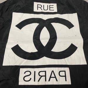 May include: Black fabric with a large white rectangle featuring a black interlocking double C logo. The word "RUE" is above the logo, and "PARIS" is below it, in white.