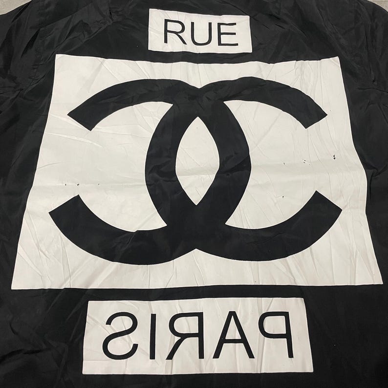 May include: Black fabric with a large white rectangle featuring a black interlocking double C logo. The word "RUE" is above the logo, and "PARIS" is below it, in white.