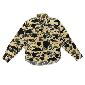 May include: A long-sleeved button-down shirt with a camouflage pattern in shades of tan, black, and green. The shirt features a classic collar, buttoned cuffs, and a curved hemline. The design includes star and ape graphics.
