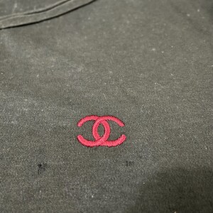 Puede incluir: Camiseta negra con un peque&ntilde;o logotipo rojo bordado en el pecho. El logotipo es un dise&ntilde;o de doble C estilizado. La camiseta tiene cuello redondo y parece estar hecha de una tela suave.