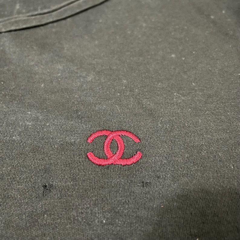 Puede incluir: Camiseta negra con un peque&ntilde;o logotipo rojo bordado en el pecho. El logotipo es un dise&ntilde;o de doble C estilizado. La camiseta tiene cuello redondo y parece estar hecha de una tela suave.