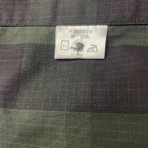 May include: Close-up of a fabric with a grid pattern in shades of gray and green. A white tag displays text and symbols, including "X 862026", "綿 100%", and Japanese characters. The fabric appears to be cotton.