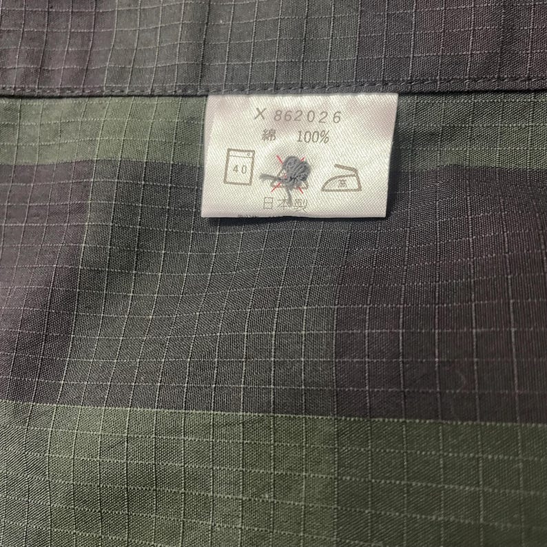 May include: Close-up of a fabric with a grid pattern in shades of gray and green. A white tag displays text and symbols, including "X 862026", "綿 100%", and Japanese characters. The fabric appears to be cotton.