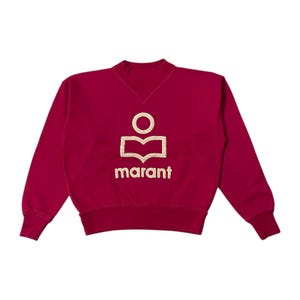 May include: A maroon sweatshirt with a crew neck and long sleeves. The front features a gold logo with the word "marant" below it. The logo is a stylized design of a person with an open book.