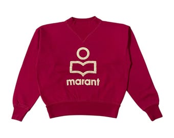 May include: A maroon sweatshirt with a crew neck and long sleeves. The front features a gold logo with the word "marant" below it. The logo is a stylized design of a person with an open book.