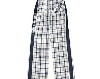 May include: White trousers featuring a navy blue and black plaid pattern. The pants have a wide-leg silhouette with a dark vertical stripe down each side. The text CHRISTIAN DIOR is printed in white on the right side stripe, and a small branded tag hangs from the waist.