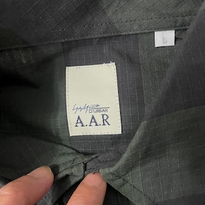 May include: Close-up of a dark green button-down shirt with a textured pattern. The shirt features a white label with the text "A.A.R" and "Durban" printed on it. A size "L" tag is visible.