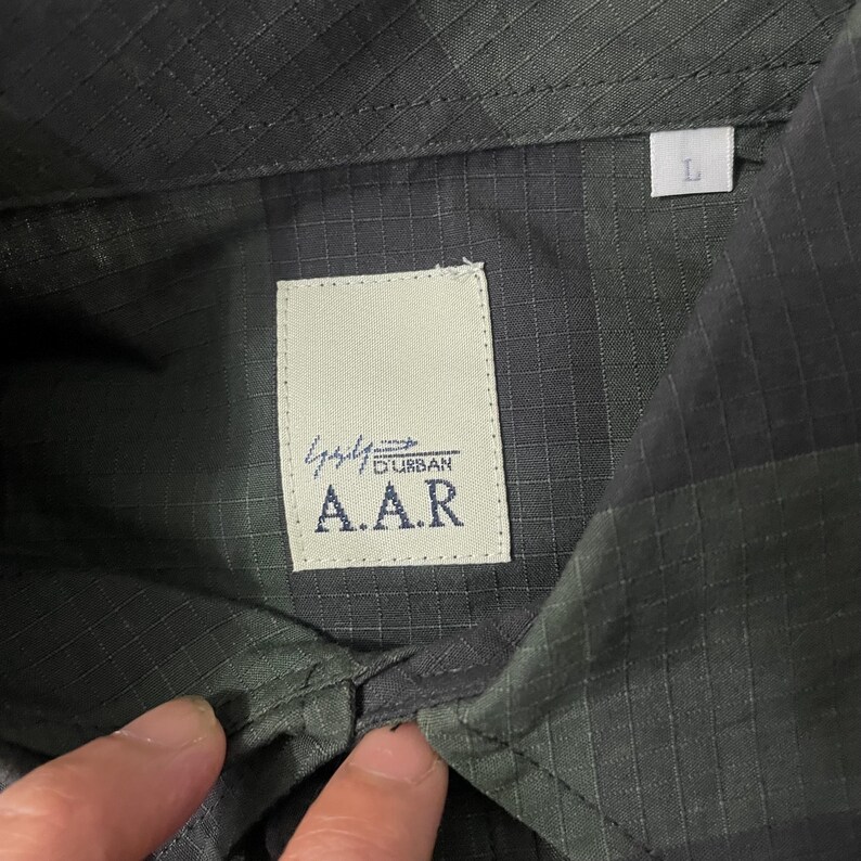 May include: Close-up of a dark green button-down shirt with a textured pattern. The shirt features a white label with the text "A.A.R" and "Durban" printed on it. A size "L" tag is visible.