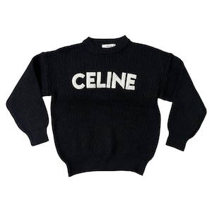 Puede incluir: Suéter de punto negro con la palabra "CELINE" en letras blancas grandes en el pecho. El suéter tiene una textura acanalada y mangas largas. El cuello es redondo. El nombre de la marca también es visible en la parte posterior del cuello.
