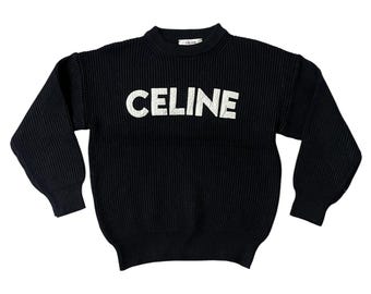 May include: Black knit sweater with the word "CELINE" in large white letters across the chest. The sweater has a ribbed texture and long sleeves. The neckline is a crew neck. The brand name is also visible on the back of the neck.