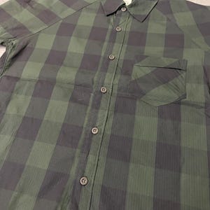 May include: Green and black plaid button-up shirt. The shirt features a classic collar, a chest pocket, and short sleeves. The fabric has a subtle sheen, and the buttons are a dark, complementary color. The plaid pattern is a large, bold design.