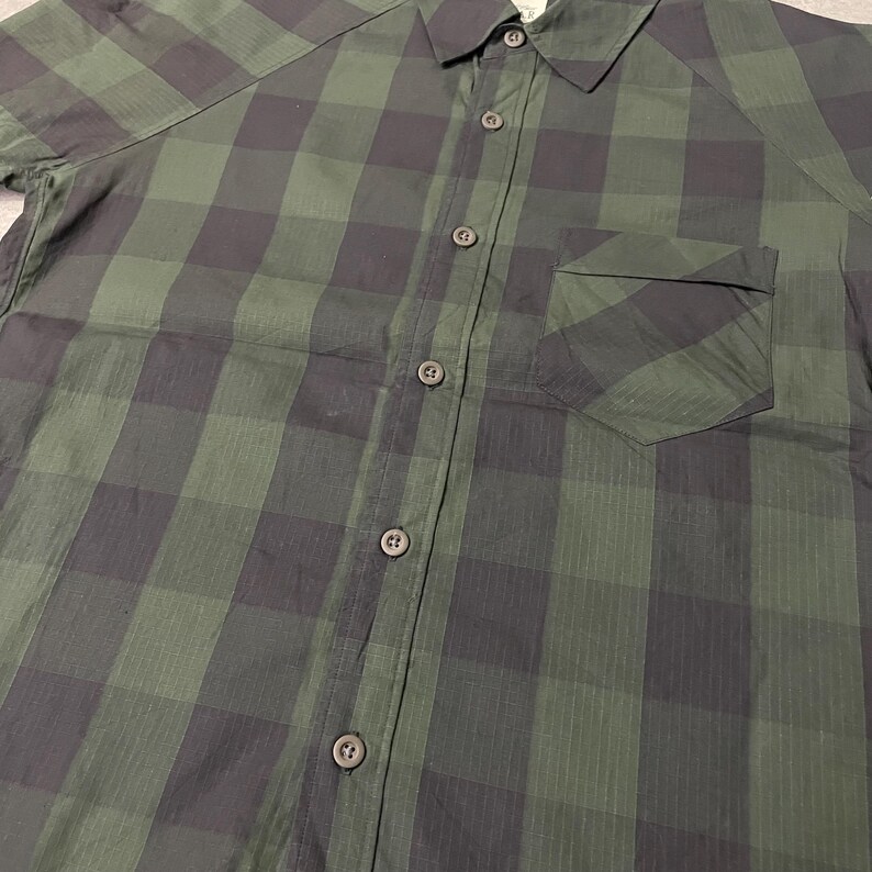May include: Green and black plaid button-up shirt. The shirt features a classic collar, a chest pocket, and short sleeves. The fabric has a subtle sheen, and the buttons are a dark, complementary color. The plaid pattern is a large, bold design.