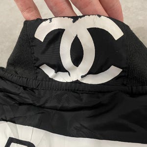 May include: Black and white garment featuring a large white logo. The logo is centered on a black background. The garment has a black ribbed collar and a black nylon-like material.