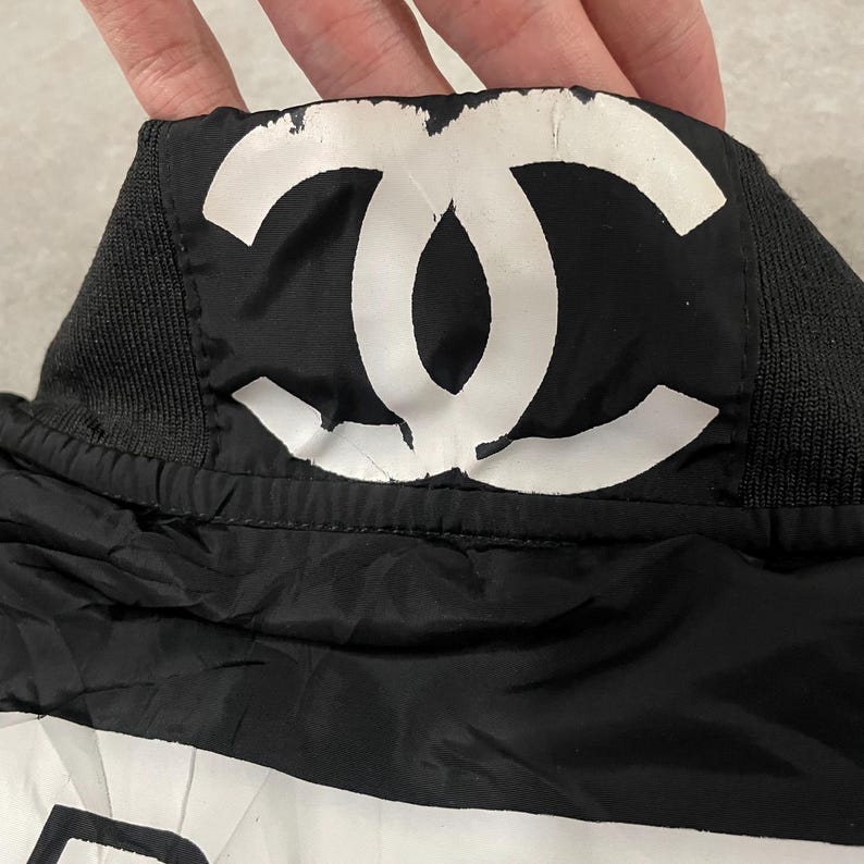 May include: Black and white garment featuring a large white logo. The logo is centered on a black background. The garment has a black ribbed collar and a black nylon-like material.