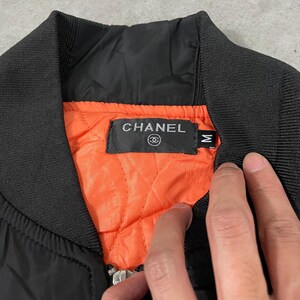 May include: Black bomber jacket with a ribbed collar and cuffs. The interior lining is bright orange and quilted. The jacket features a label with the brand name "CHANEL" in silver lettering.