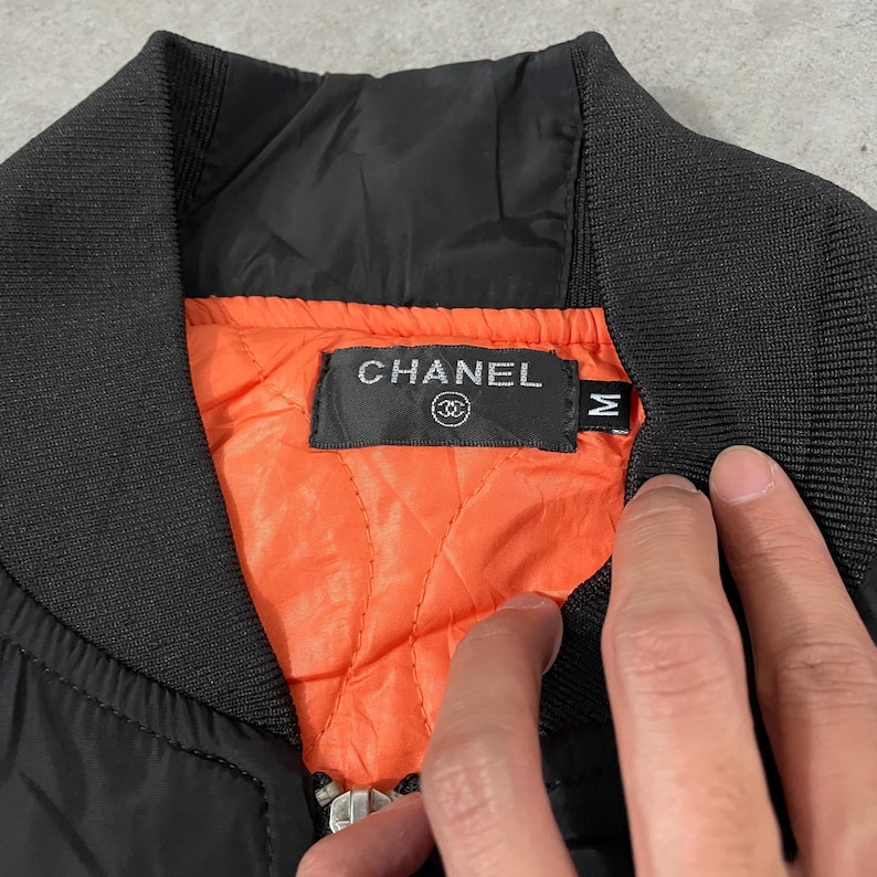 May include: Black bomber jacket with a ribbed collar and cuffs. The interior lining is bright orange and quilted. The jacket features a label with the brand name "CHANEL" in silver lettering.