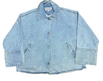 May include: A light blue denim jacket featuring a pointed collar, long sleeves with wide cuffs, and a front button closure. The interior label displays the text LOEWE. The jacket has two side pockets and a light wash finish throughout the fabric.