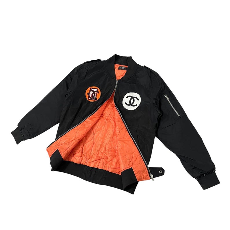 May include: Black bomber jacket with orange lining. Features a white and black logo patch on the chest and an orange and black logo patch on the upper left sleeve. Includes a zipper closure and a small pocket on the left sleeve.
